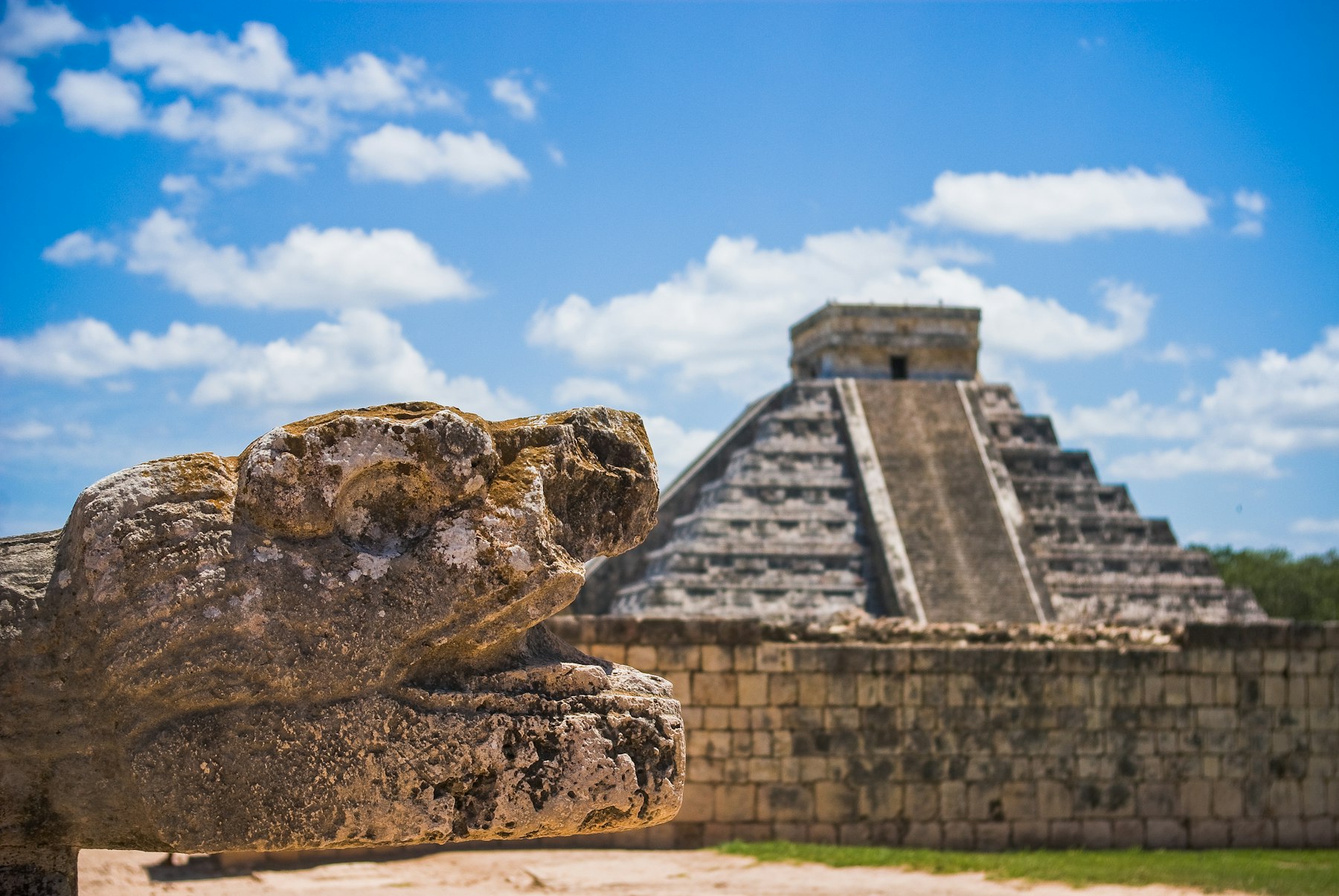 A carved Maya serpent head sculpture in the foreground with the El Castillo pyramid of Chichén Itzá rising under a blue sky behind it.