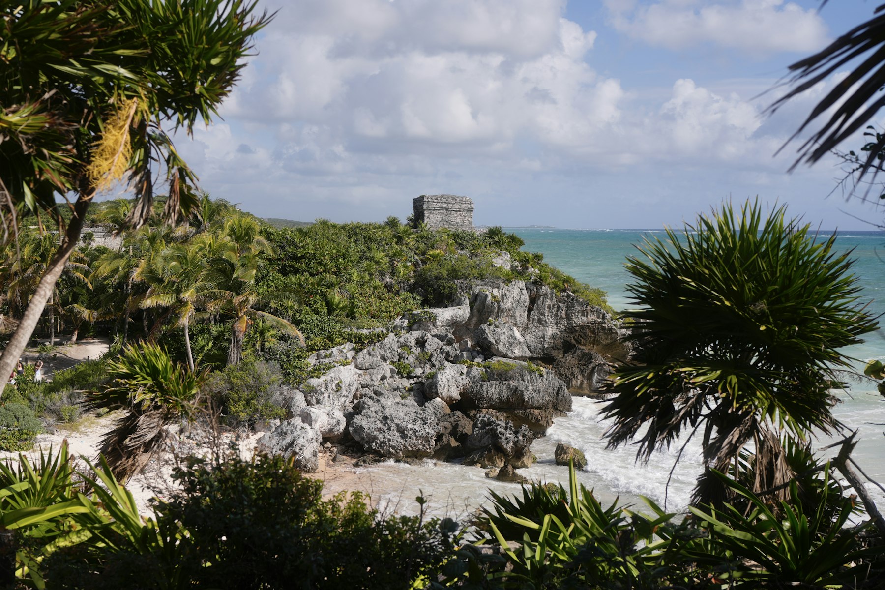 A stone Maya watchtower at Tulum perched on a coastal cliff above turquoise Caribbean water, framed by palms.